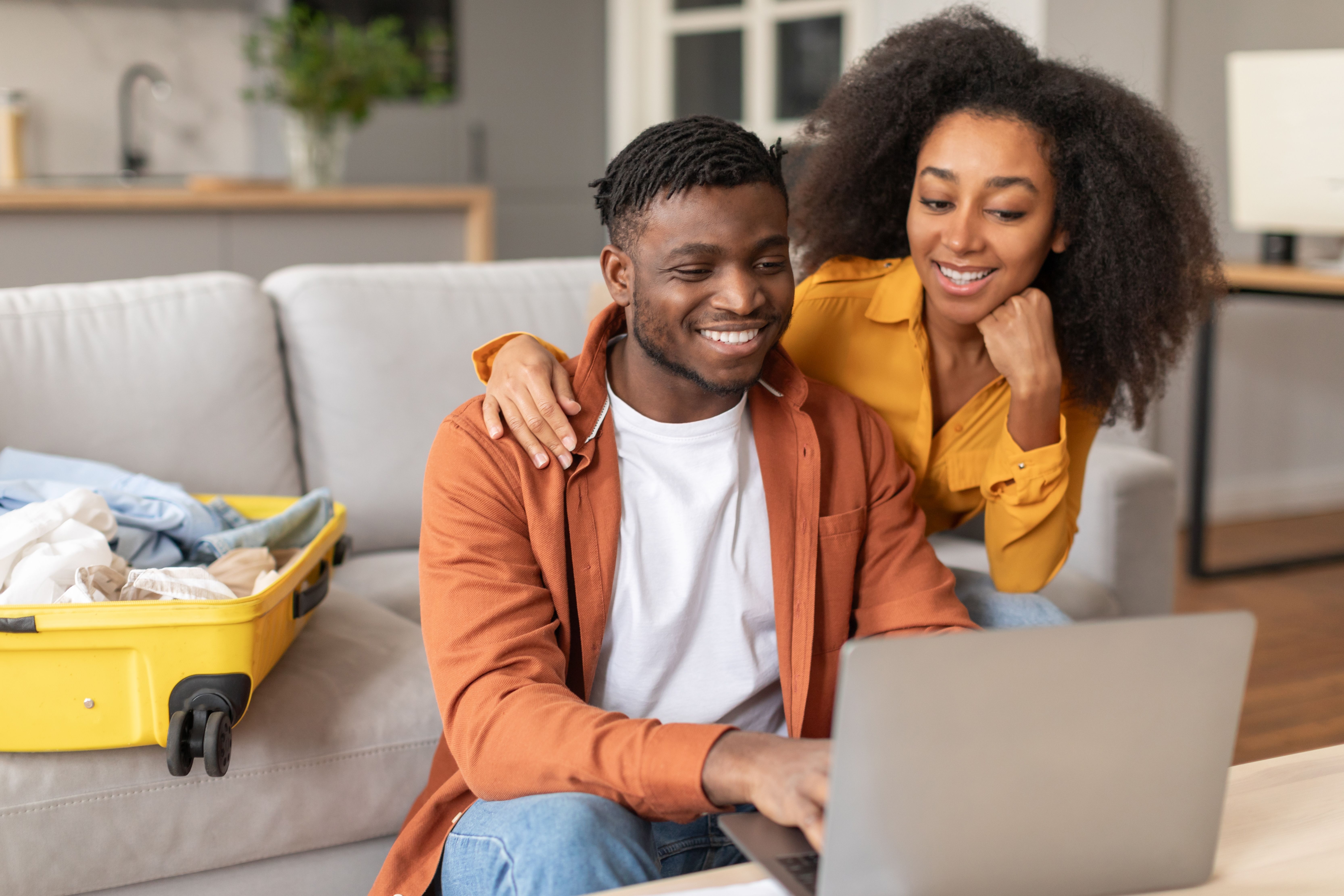 Smiling couple researching and booking their next cruise vacation on a laptop at home, with an open suitcase beside them as they prepare for their trip.