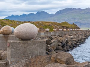 Dj&uacute;pivogur, Island &ndash; Reihe von Eiform-Skulpturen auf Betonsockeln am Ufer mit Bergen im Hintergrund