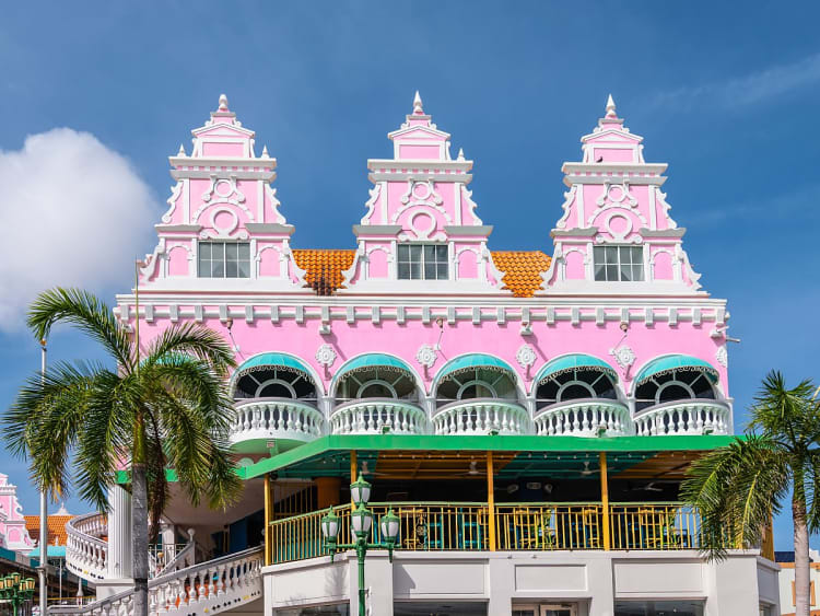 Oranjestad, Aruba &ndash; rosa Giebelhaus mit verspielter Fassade und Palmen vor blauem Himmel