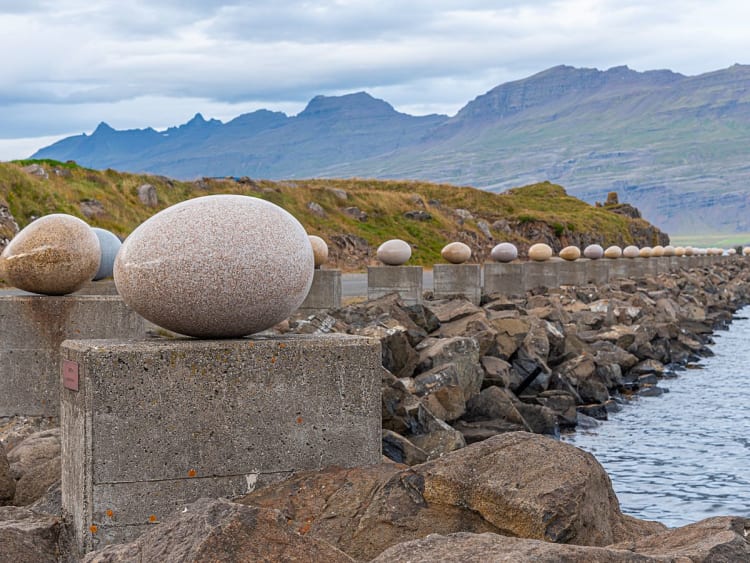 Dj&uacute;pivogur, Island &ndash; Reihe von Eiform-Skulpturen auf Betonsockeln am Ufer mit Bergen im Hintergrund