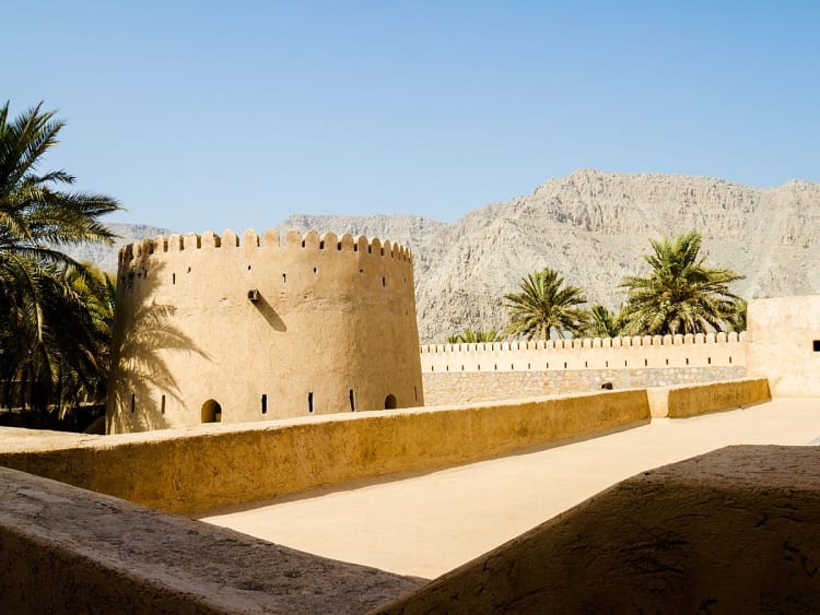 Khasab, Oman &ndash; Historische Festung mit Rundturm, umgeben von Palmen und Bergen im Hintergrund