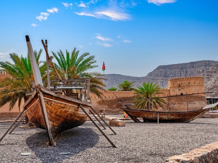 Khasab, Oman &ndash; Traditionelle Holzboote liegen vor der alten Festung mit Palmen und Bergen