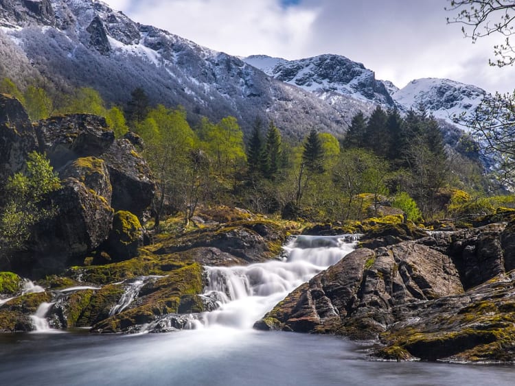 Rosendal, Norwegen - Wasserfall flie&szlig;t &uuml;ber Felsen vor schneebedeckten Bergen