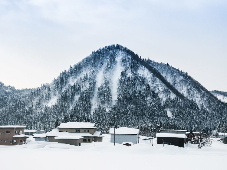 Sakata, Japan - Verschneites Dorf vor bewaldetem Berg unter grauem Winterhimmel