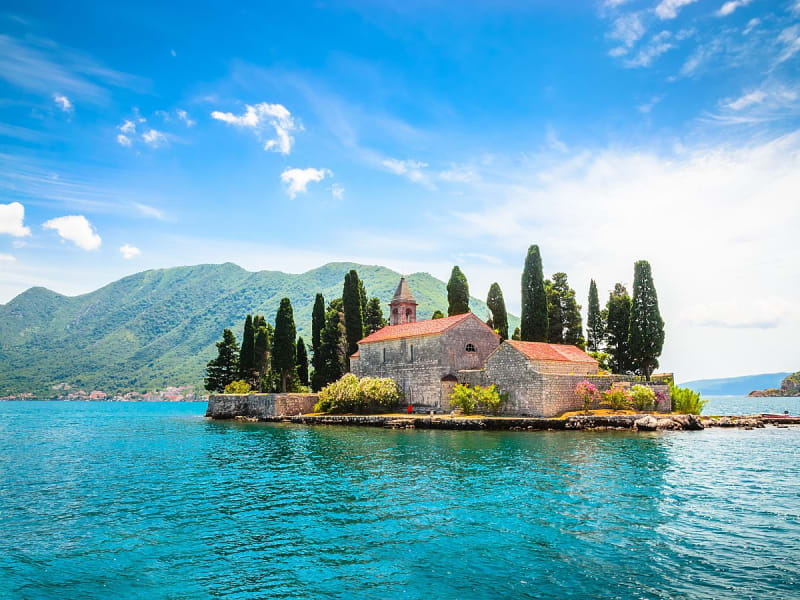 Kleine Klosterinsel mit Zypressen im t&uuml;rkisblauen Wasser vor gr&uuml;ner Bergkulisse.