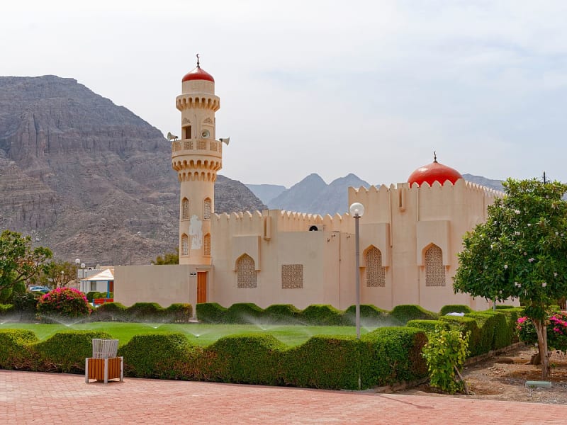Khasab, Oman &ndash; Wei&szlig;e Moschee mit roter Kuppel und gepflegtem Garten vor Gebirgskulisse