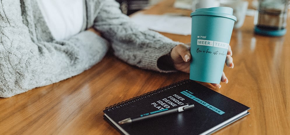 Mitarbeitende hält e-hoi Kaffeebecher in der Hand, ein Notizbuch und Stift liegen auf dem Tisch.