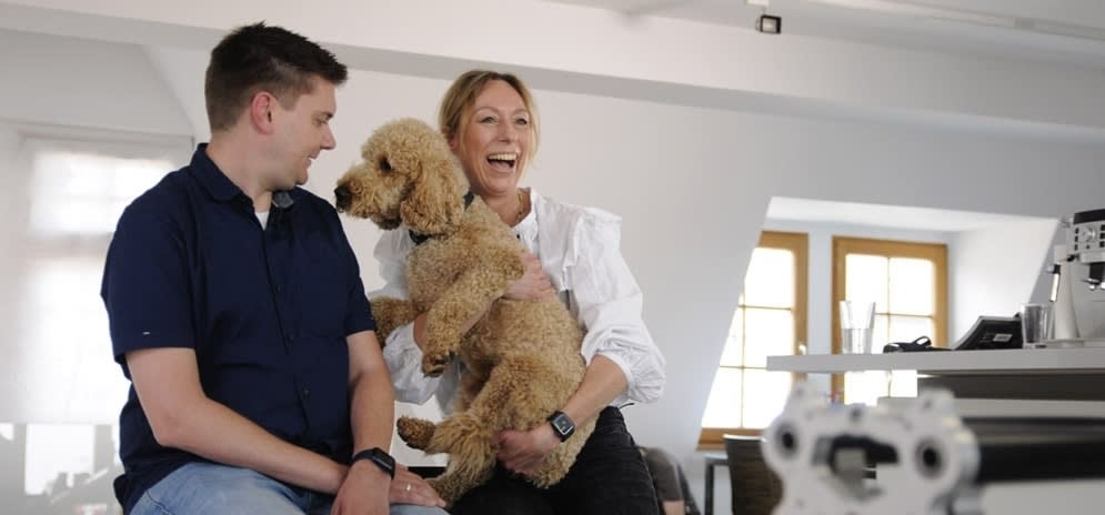Zwei Mitarbeitende lachend in Interaktion mit einem der Büro-Hunde.