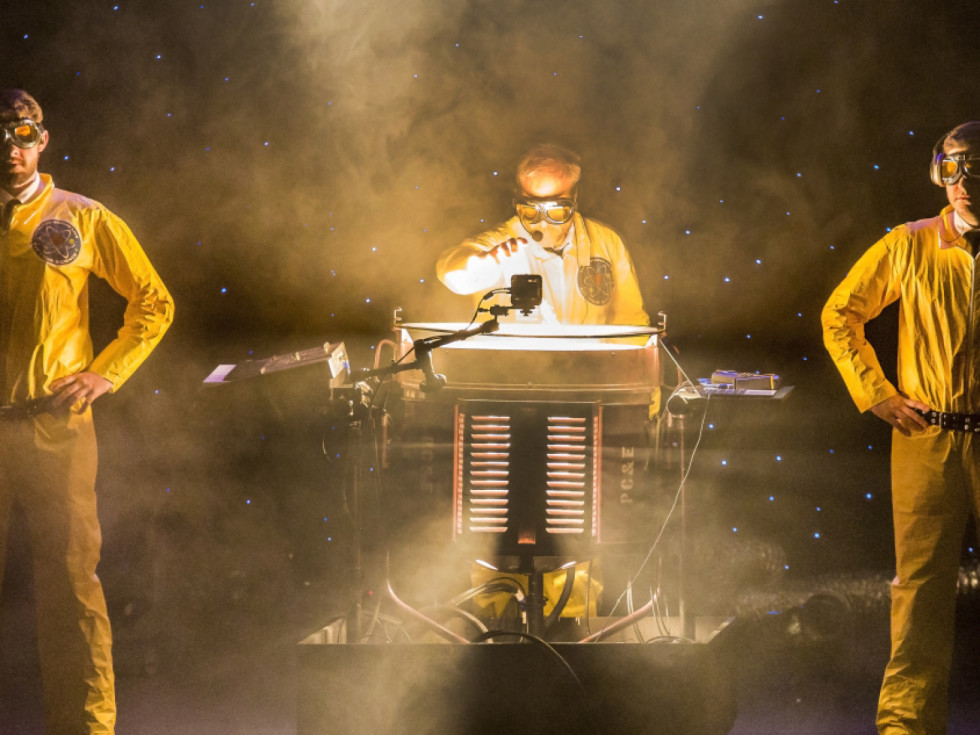 Alton Brown and two assistants stand in yellow hazmat suits around a mysterious contraption.