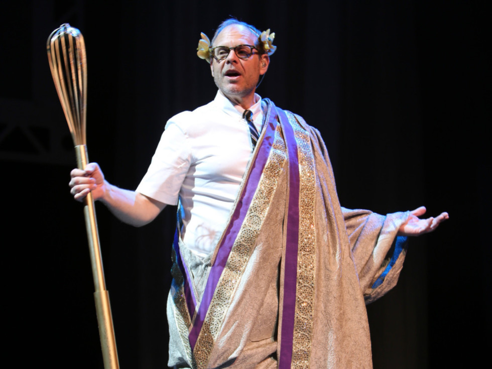 Celebrity chef and TV host Alton Brown poses onstage in a toga with a giant whisk.