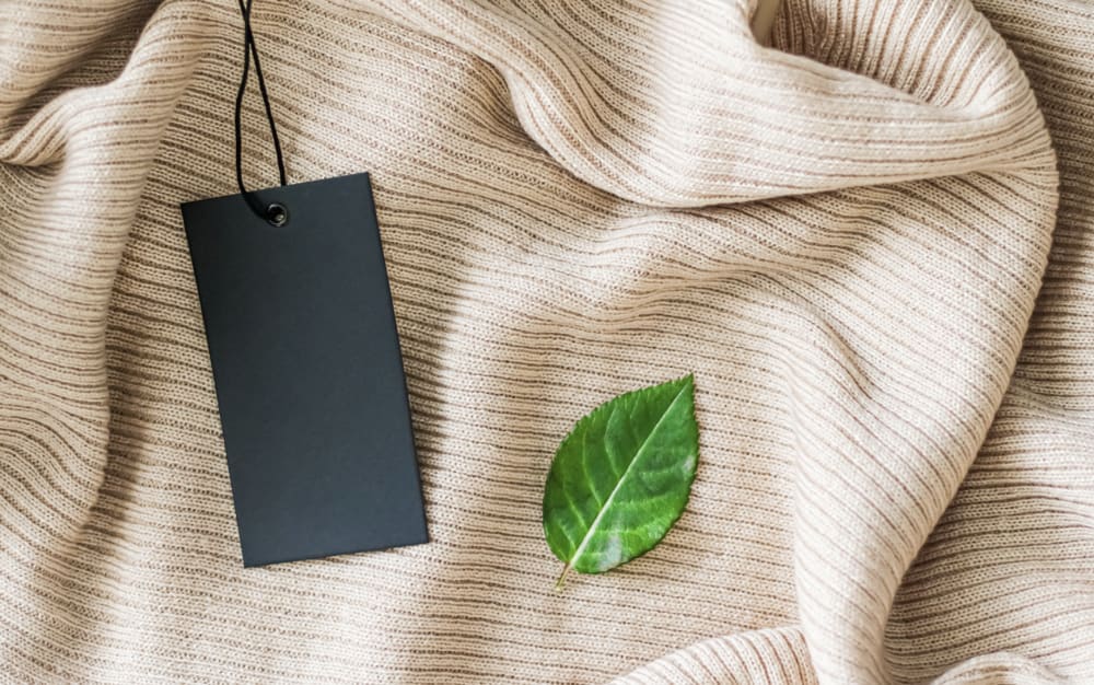 Flat laid beige woollen garment with a small black label and small green leaf laying on top