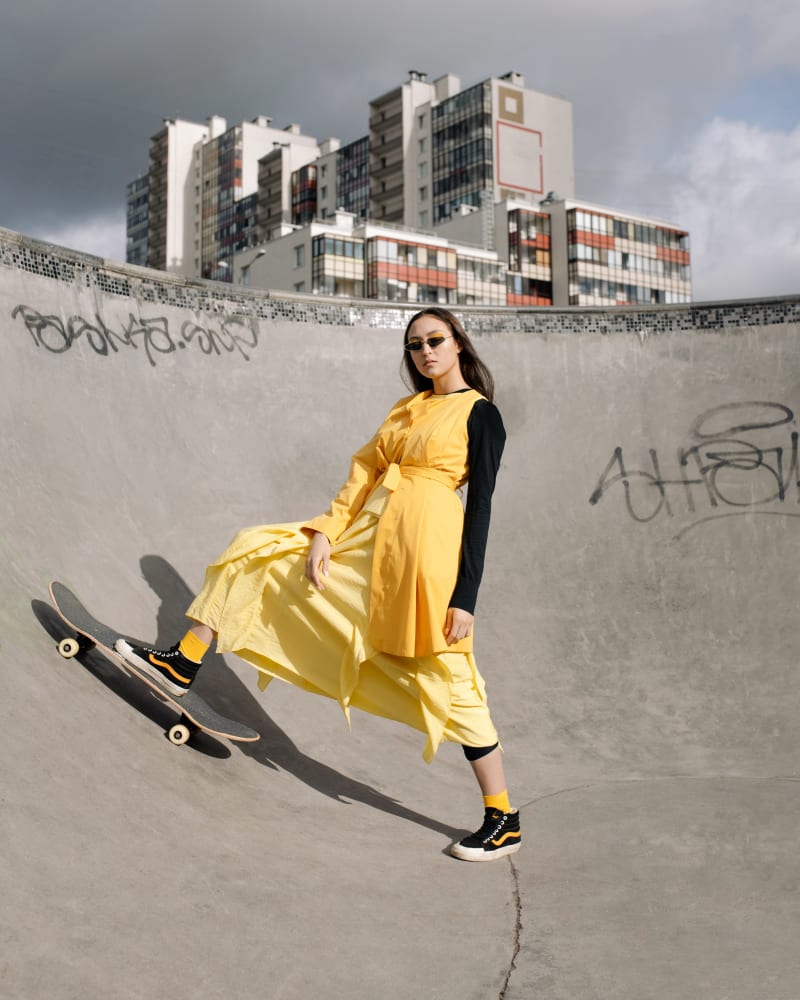 Woman in yellow dress, street wear and trainers at a skatepark, standing on a skateboard