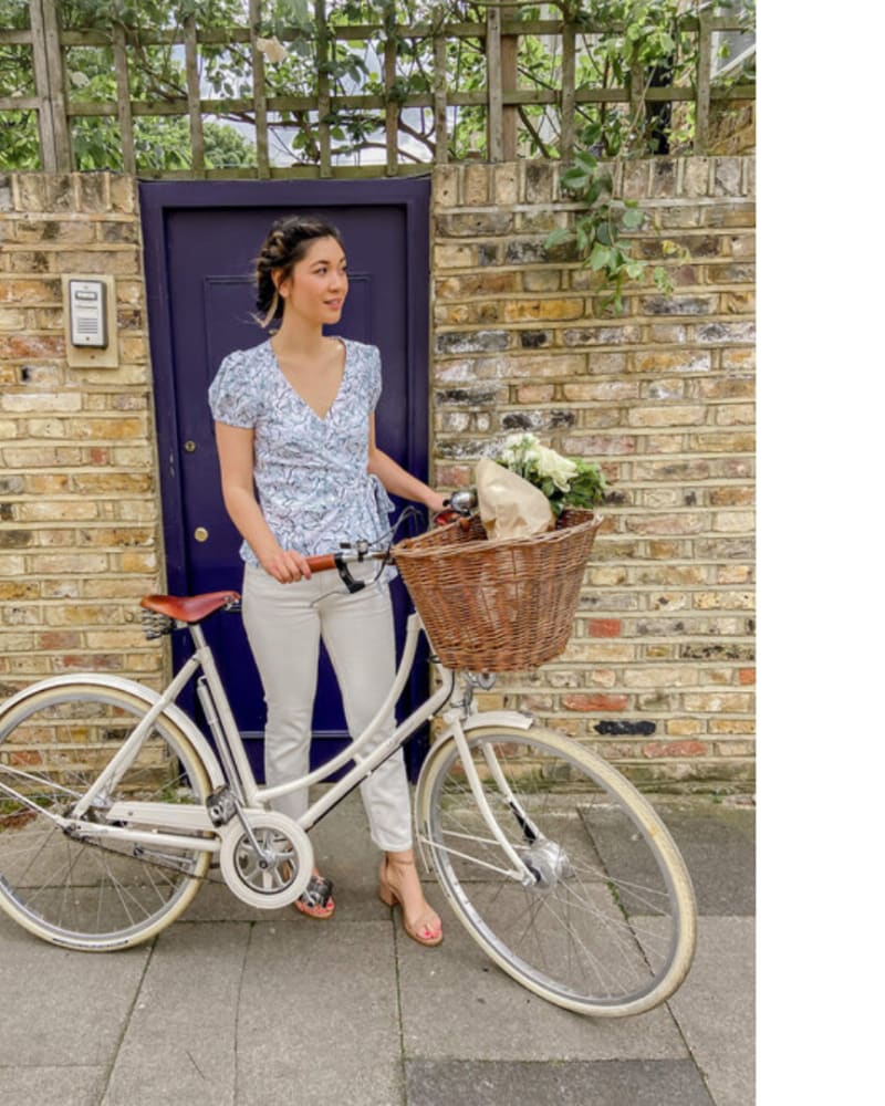 Woman standing outdoors with a white bicycle, wearing a light coloured v-neck wrap top in short sleeves.