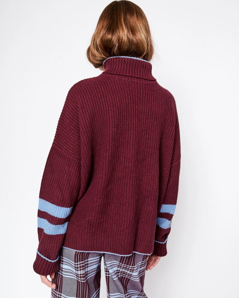 A girl facing away from the camera wearing a burgundy chunky knit turtle neck jumper with blue horizontal stripes at the end of the sleeves