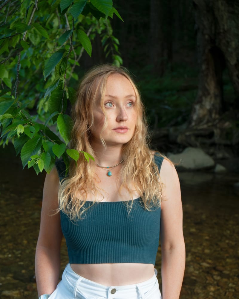 Woman standing outside in a wood wearing a green ribbed square neck crop top and white trousers. 