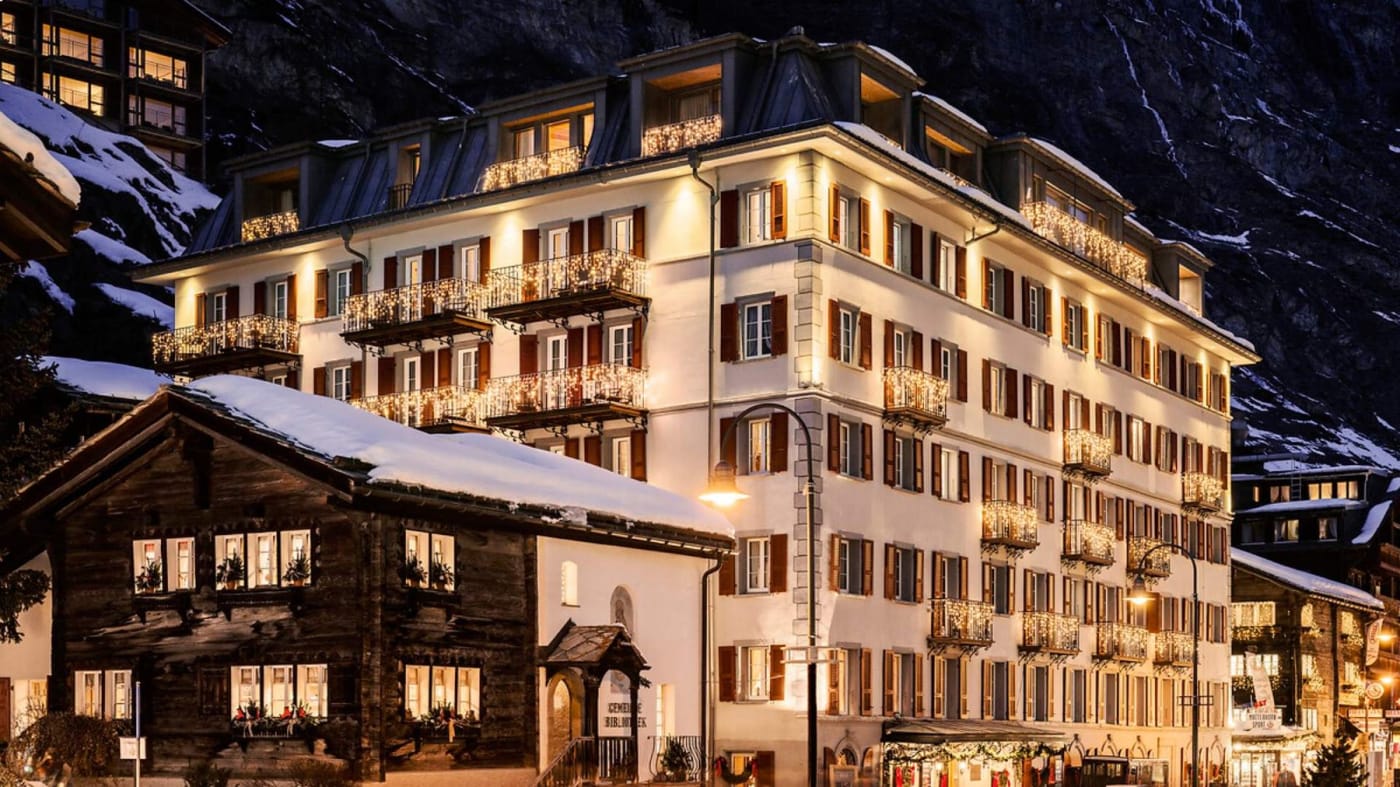 Die beleuchtete Fassade des Hotel Monte Rosa an der Bahnhofstrasse in Zermatt an einem Winterabend, neben einem alten Holzchalet