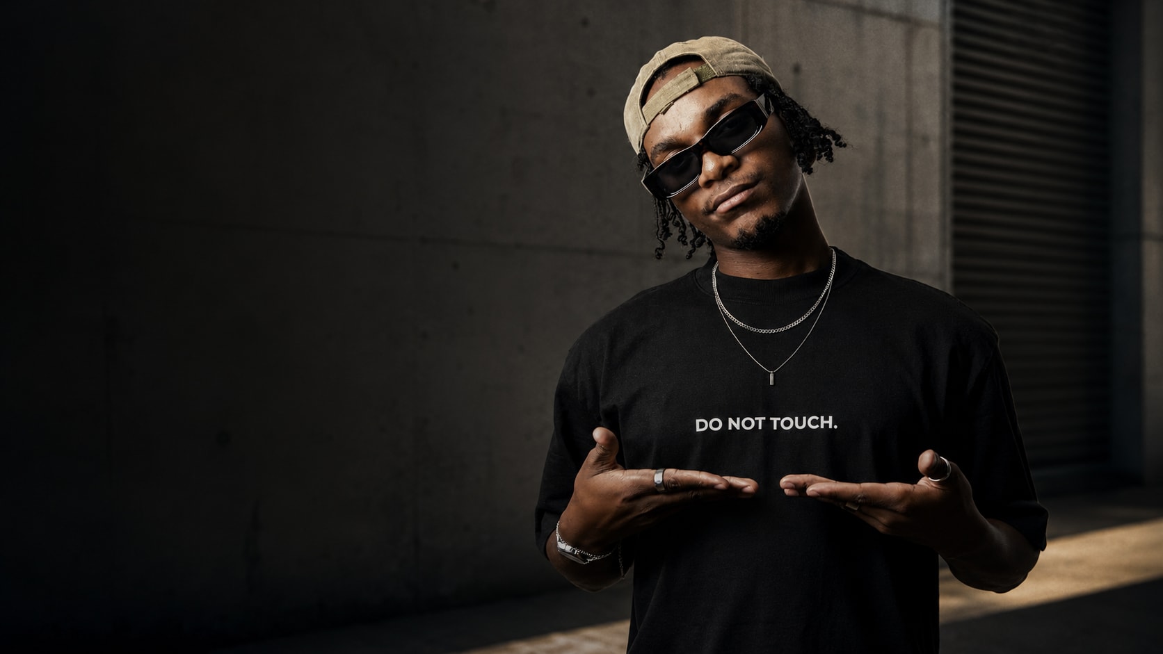 Do Not Touch model in sunglasses presenting the wordmark on an oversized black T-shirt against a concrete wall