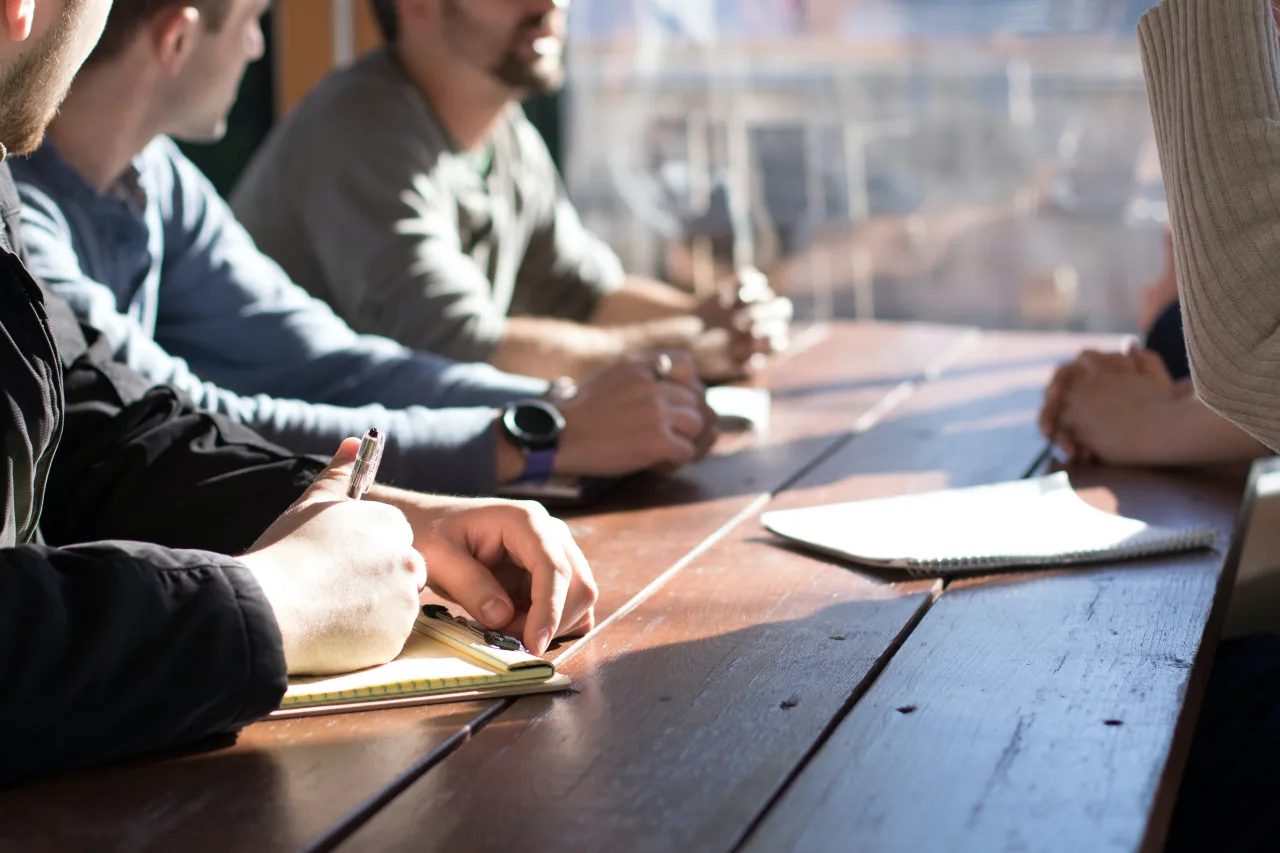 Group of people engaged in a discussion at a sunlit wooden table, with focus on hands taking notes and holding pens, suggesting a collaborative work environment or a team meeting.