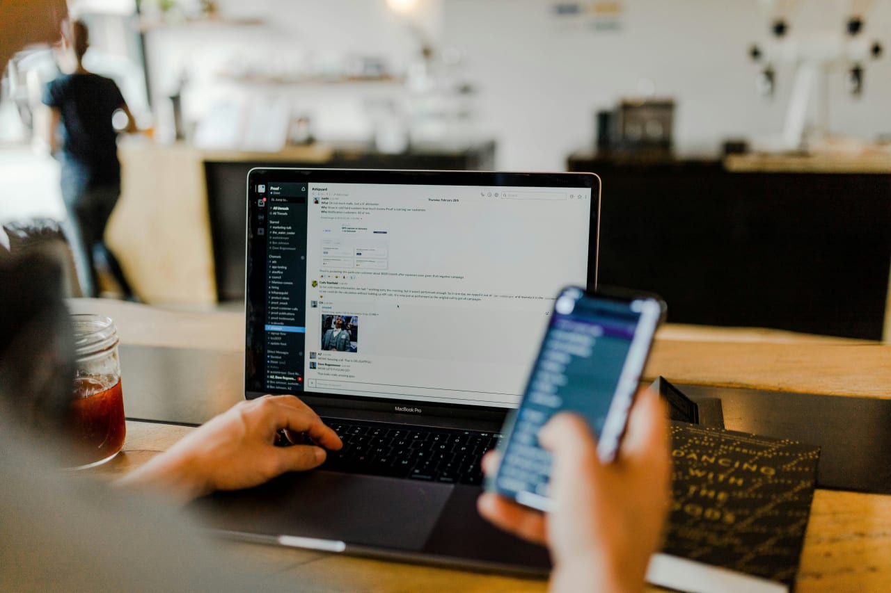 A person multitasks using Slack on a laptop and a mobile phone in a cafe setting, managing communications across multiple devices.