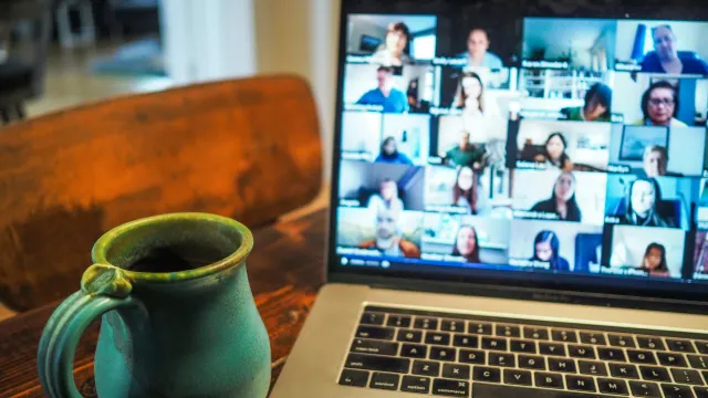 A laptop screen displaying a virtual meeting with multiple participants, accompanied by a ceramic mug on a wooden table in the foreground, depicts the new normal of remote collaboration.
