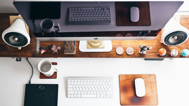 Overhead view of a modern, minimalist workspace featuring an Apple monitor, keyboard, and mouse on a wooden desk.