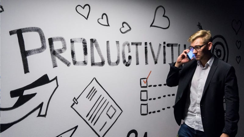 A man in a business suit is talking on a smartphone, standing against a wall with a graffiti-style drawing that spells 'PRODUCTIVITY' and doodles of checklists and targets.