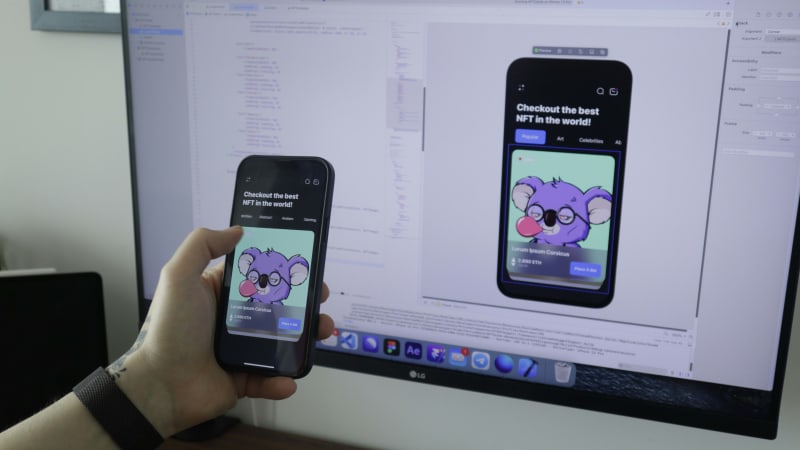 A developer testing an iOS app featuring an NFT on a smartphone, with the app's code and user interface displayed on the computer screen in the background.