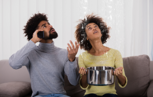 property management image: young couple staring at ceiling with water leaking; the man is on the phone while the woman holds a pot to catch the water leaking