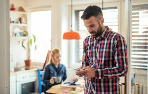 mobile rent payments image: picture of a well-groomed man wearing flannel on his mobile phone, blond woman drinking coffee and reading newspaper in the background