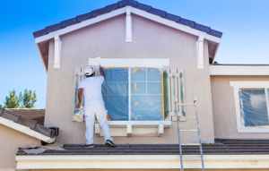 ultimate guide to house flipping header image: man in white shirt and pants painting tan house