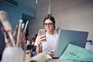 lease signing: woman with hair in ponytail and glasses smiling at phone sitting next to laptop