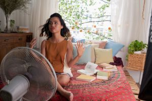 keep a room cool without ac hero image: young woman sitting in bed in front of fan