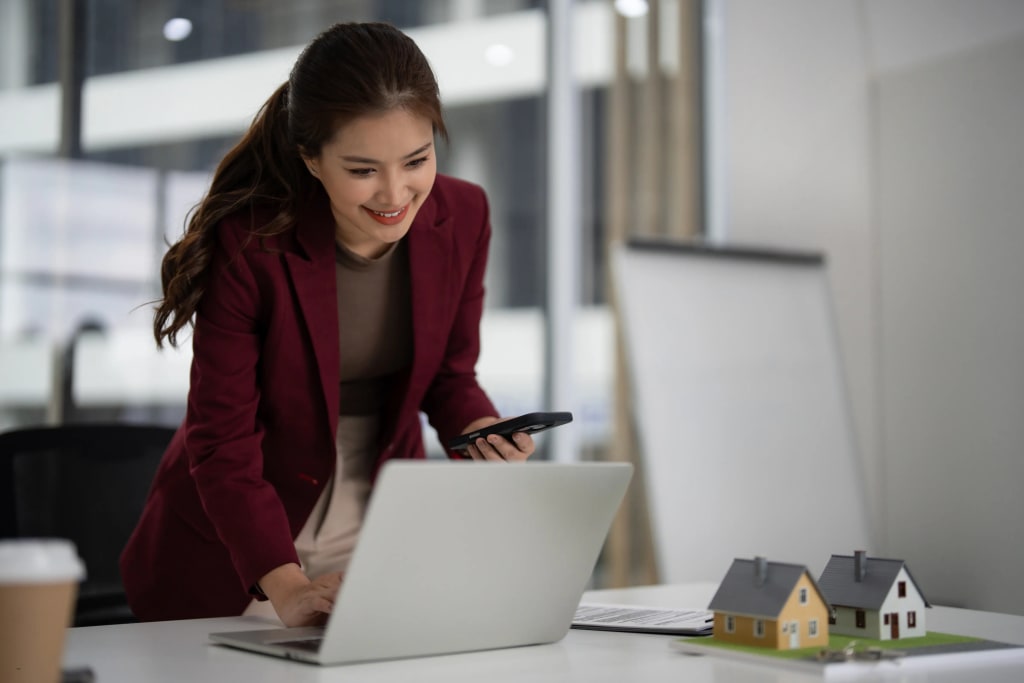 Property management app in use by real estate professional working on laptop while checking mobile alerts beside miniature house models.