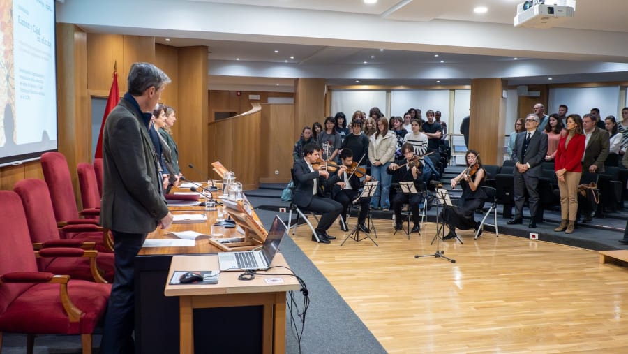 A formal event featuring a group of musicians playing string instruments in front of an audience, with several speakers at a table and a presentation displayed in the background.