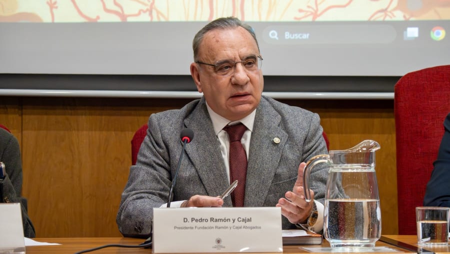 D. Pedro Ramón y Cajal, Presidente de la Fundación Ramón y Cajal Abogados, participando en una mesa redonda. En la mesa hay un vaso de agua y una jarra. Al fondo, una presentación visual.