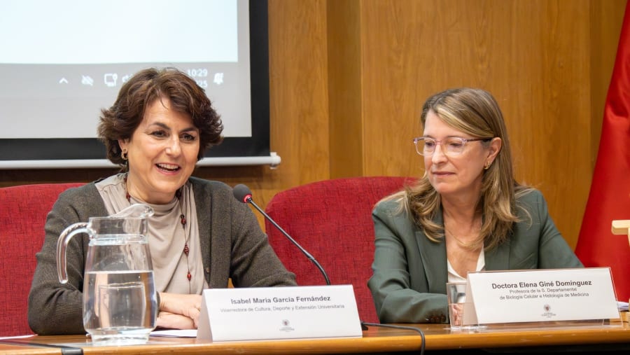 Two women sitting at a table during a conference, with a pitcher of water and name plates visible in front of them. One woman is smiling while the other appears thoughtful.