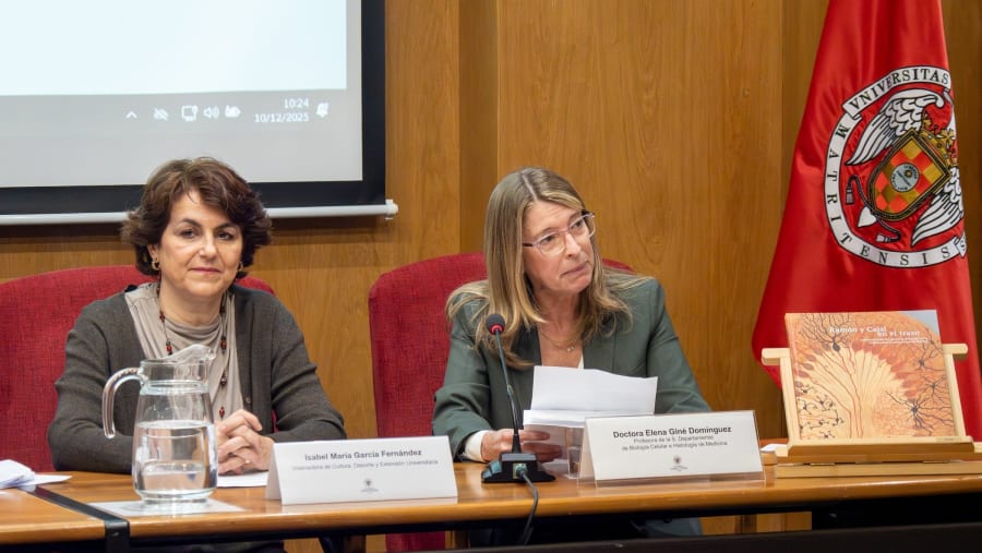 Two women sitting at a table during a presentation, with a glass pitcher of water in front of them and a book displayed on an easel beside them. One woman is reading from a document while the other is listening attentively. A university emblem is visible in the background.