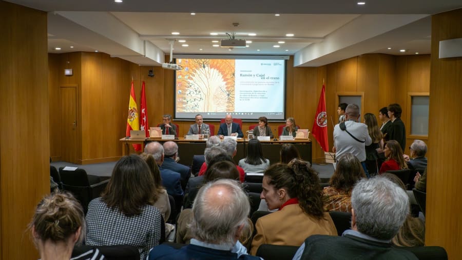 A conference scene featuring five speakers at a table, with the audience visible in the foreground. The backdrop displays artwork and official flags.