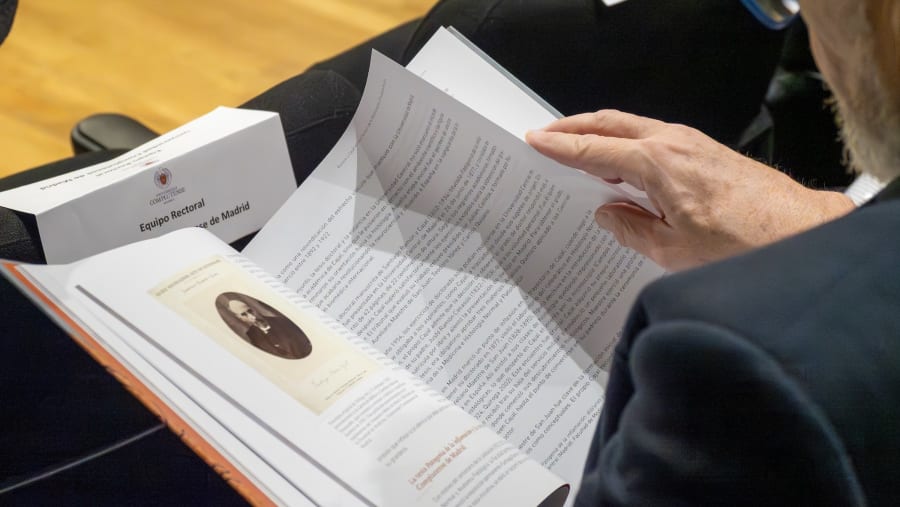 A person is holding open a document that features text, with a portrait visible. In the background, a sign reads 