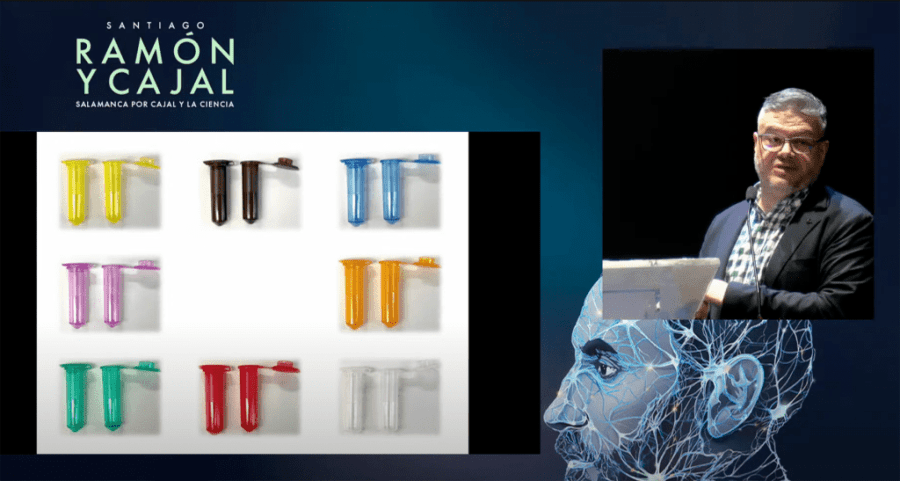 A presentation featuring colorful laboratory test tubes arranged in pairs on a white background, alongside a speaker at a podium with a digital backdrop of Santiago Ramón y Cajal