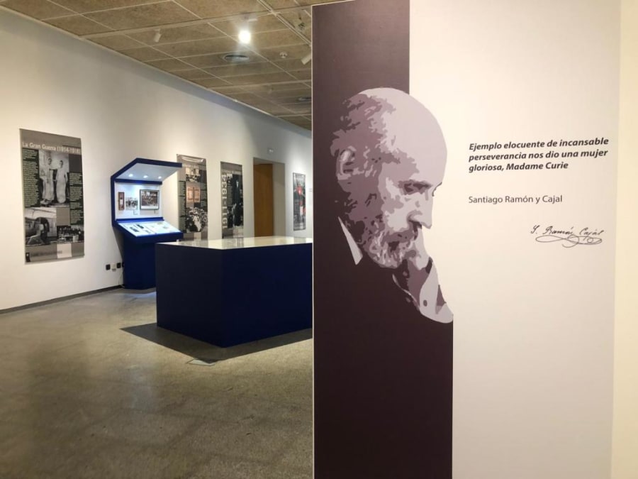 Sala de exposición sobre María Sklodowska-Curie con paneles informativos y un retrato de Santiago Ramón y Cajal, destacando su frase sobre la perseverancia de Madame Curie.
