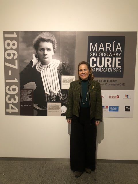 Una mujer posando frente a un panel informativo sobre María Sklodowska-Curie en una exposición, con fotografías y textos explicativos de su vida y obra.