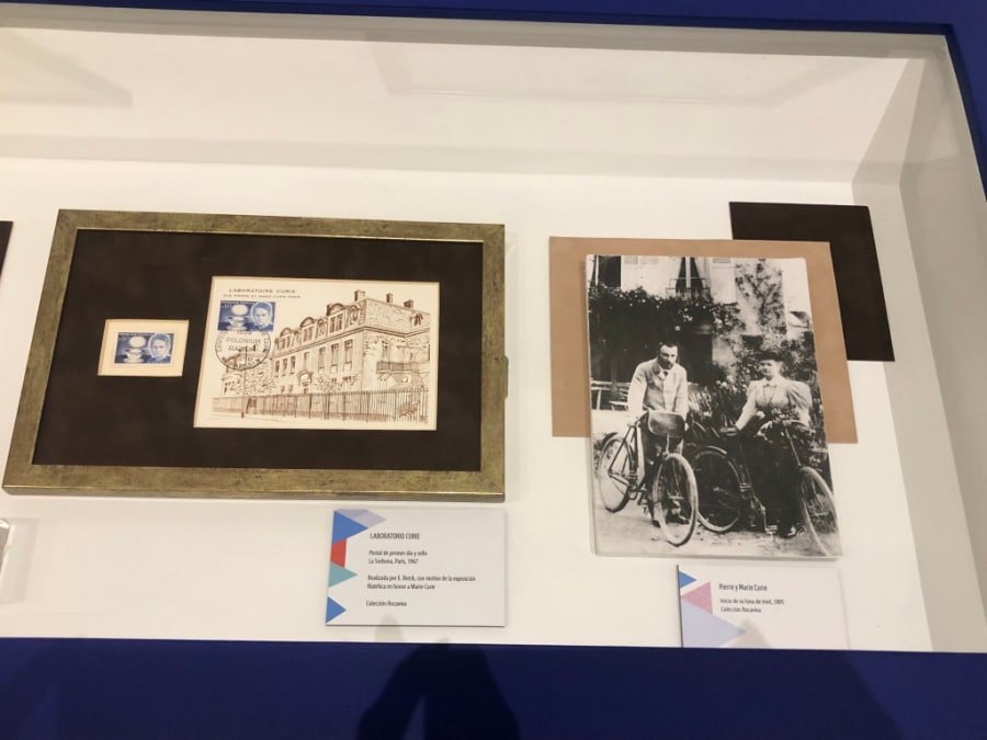 Exhibición de una carta y un sello conmemorativo del laboratorio Curie, junto a una fotografía de Pierre y Marie Curie en bicicleta, en una vitrina de la exposición sobre María Sklodowska-Curie.