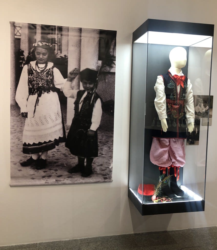 Una fotografía en blanco y negro de dos niños con trajes tradicionales, uno de ellos sosteniendo una rama, junto a un vitrina que exhibe un traje tradicional polaco adornado con medallas y accesorios culturales.