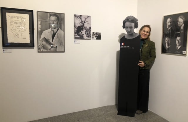 Persona posando junto a una figura de María Sklodowska-Curie en una exposición, con varias fotografías y un retrato en la pared.