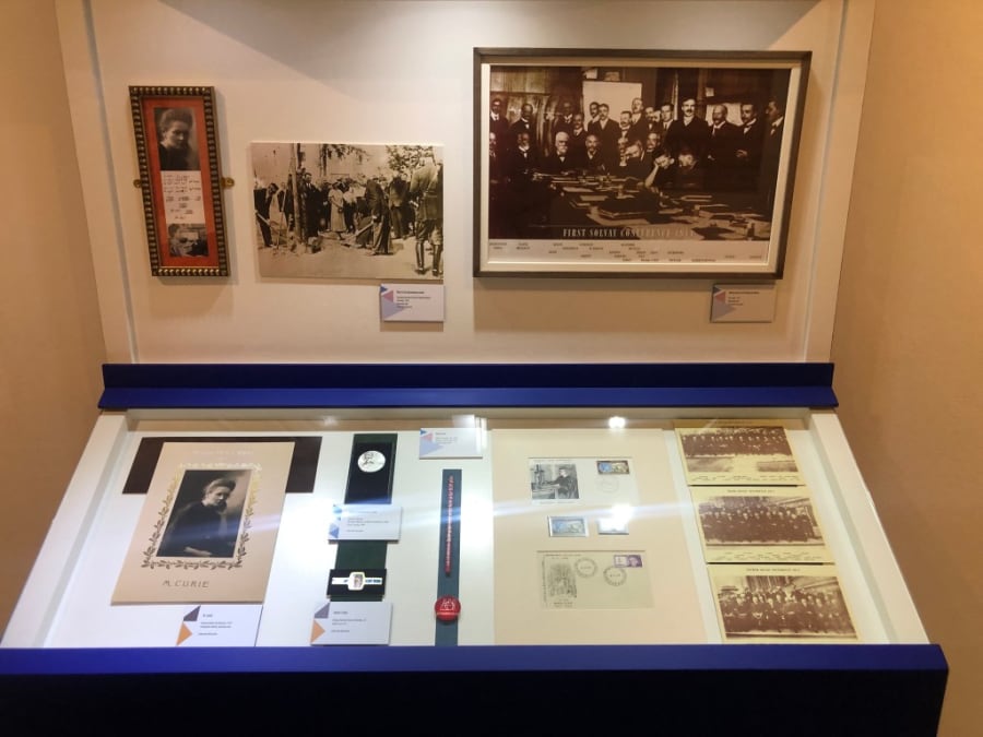 Vitrina de exposición que muestra un collage de documentos relacionados con María Sklodowska-Curie, incluyendo fotos, sellos, y un retrato enmarcado de la científica.