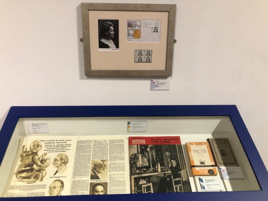 Exhibición de documentos y fotografías relacionados con María Sklodowska-Curie, incluyendo un retrato, sellos y revistas científicas, en una vitrina en la exposición sobre su vida y trabajo.
