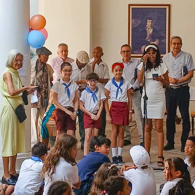 Un grupo de niños y adultos en un evento educativo, con decoraciones de fiesta y una mujer hablando en un micrófono.