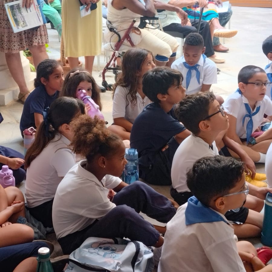 Grupo de niños sentados en un evento, algunos bebiendo de botellas de agua, mientras observan hacia adelante. Visten camisetas blancas y otras de diferentes colores, mostrando interés en la actividad.