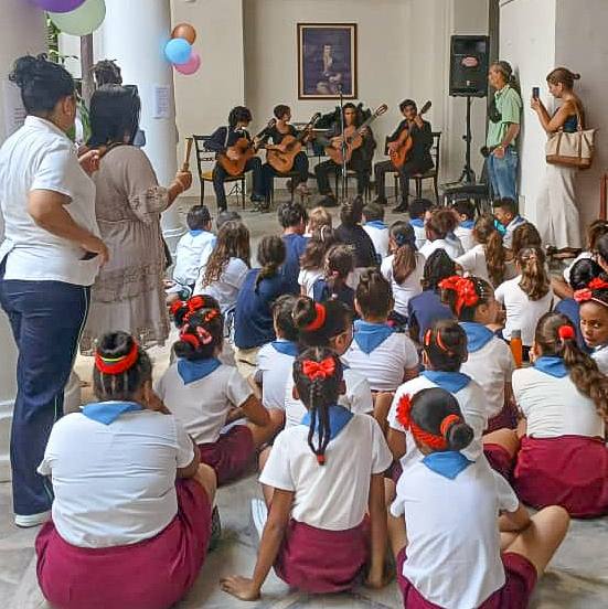 Un grupo de jóvenes músicos toca guitarras en un evento, mientras un público de niños en uniforme escolar los observa atentamente. Hay decoraciones de globos en el fondo y se puede ver a varios adultos entre el público.